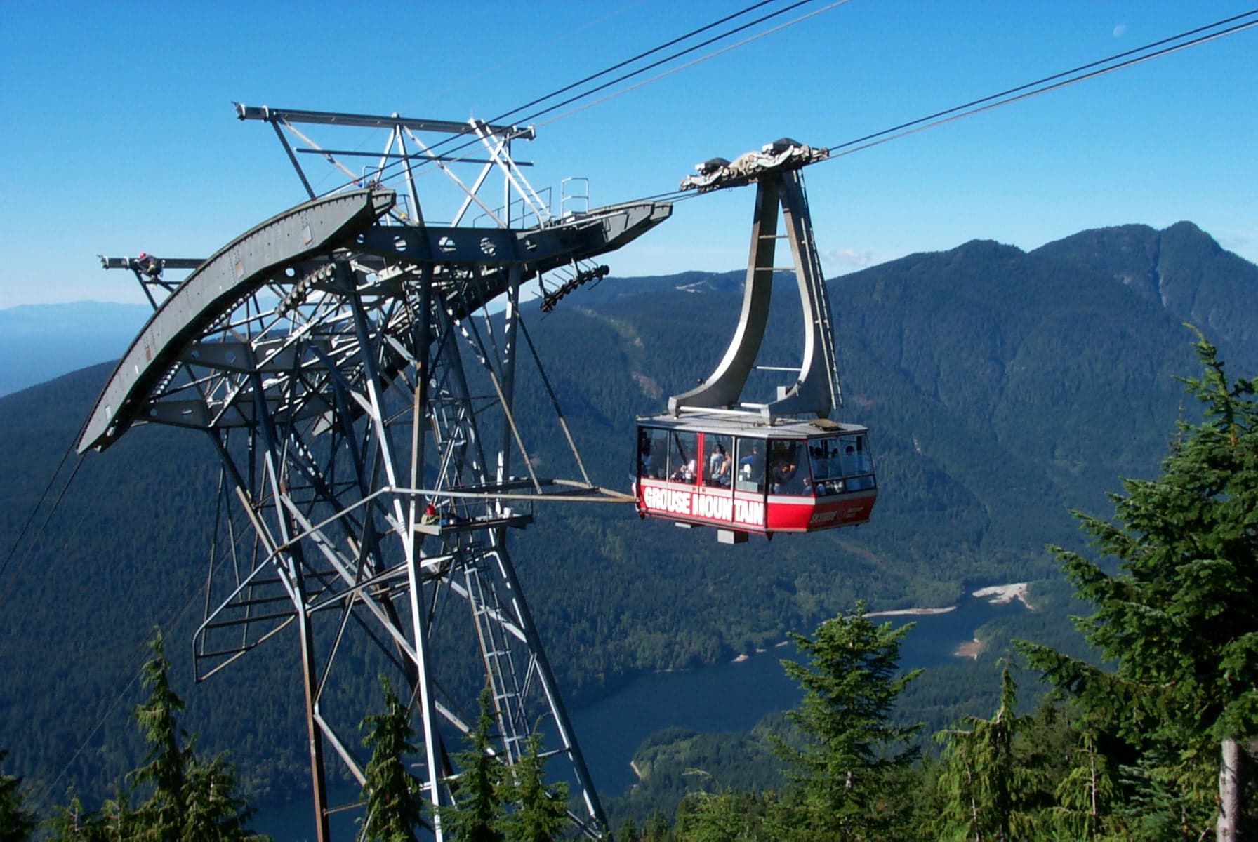 A red Grouse Mountain Skyride gondola ascending toward the summit station, with forested slopes visible below and the mountain above.