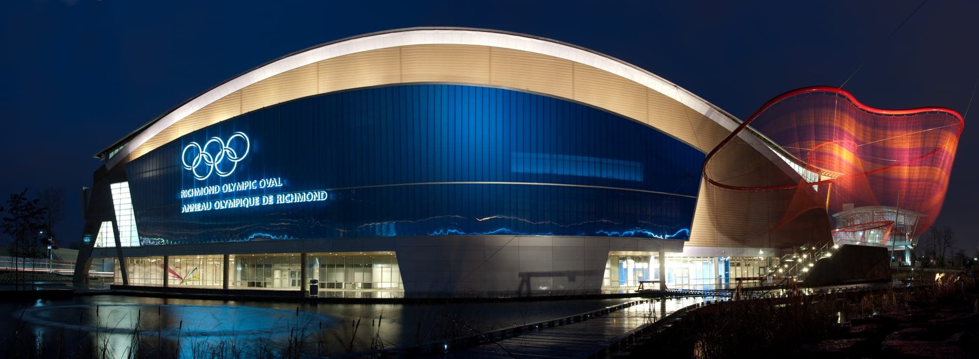 The Richmond Olympic Oval speed-skating venue at night, with its distinctive wave-form wooden roof lit up and reflections on the river.