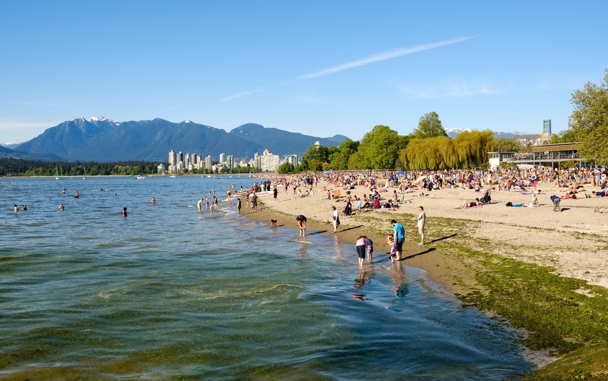 Kitsilano Beach in Vancouver with people on the sand and the downtown skyline and North Shore mountains visible across English Bay.